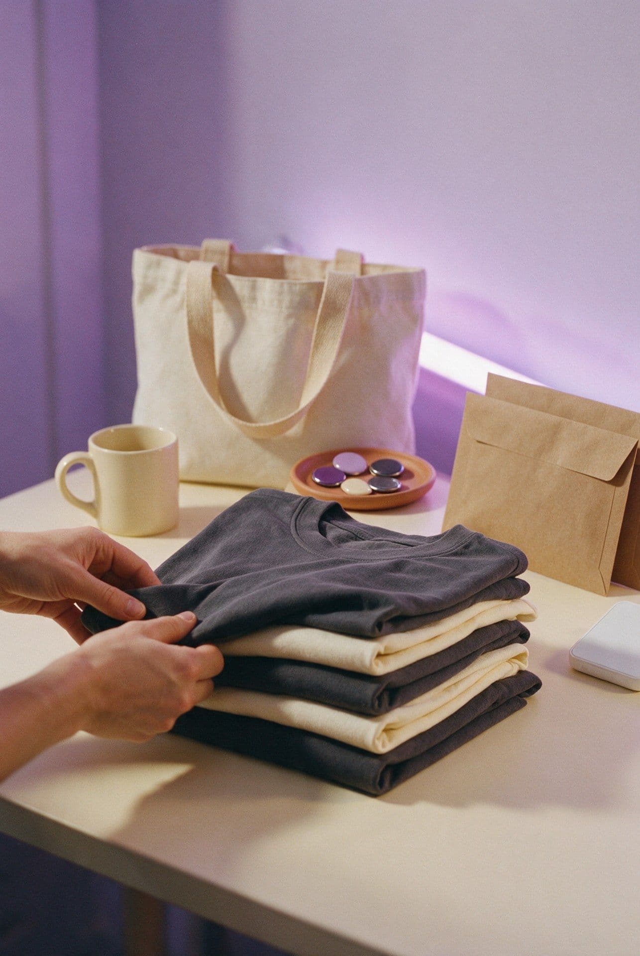 Network merchandise being packed by hand — folded shirts, a tote, pins and a card reader